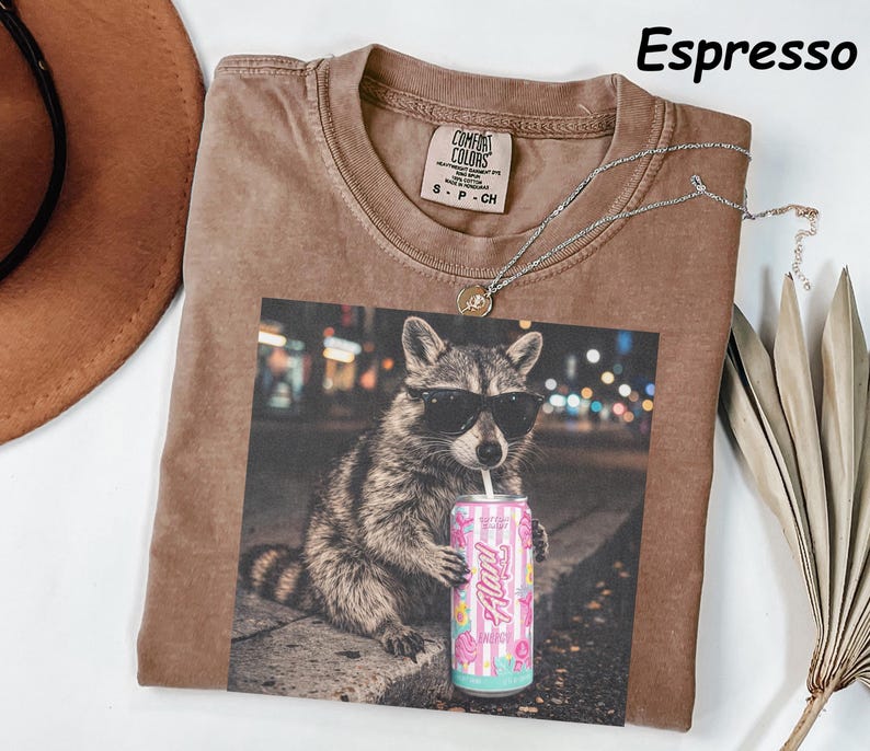 May include: Espresso-colored t-shirt featuring a graphic of a raccoon wearing sunglasses and holding a can of soda. The shirt has a crew neck and a small tag on the collar. A silver necklace is draped across the shirt.