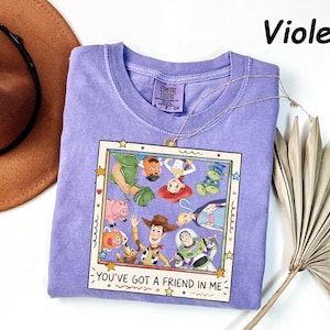 May include: A violet short-sleeved t-shirt featuring a graphic print of Toy Story characters inside a square frame. The text "YOU'VE GOT A FRIEND IN ME" is printed below the frame. The shirt has a Comfort Colors brand label at the neck.