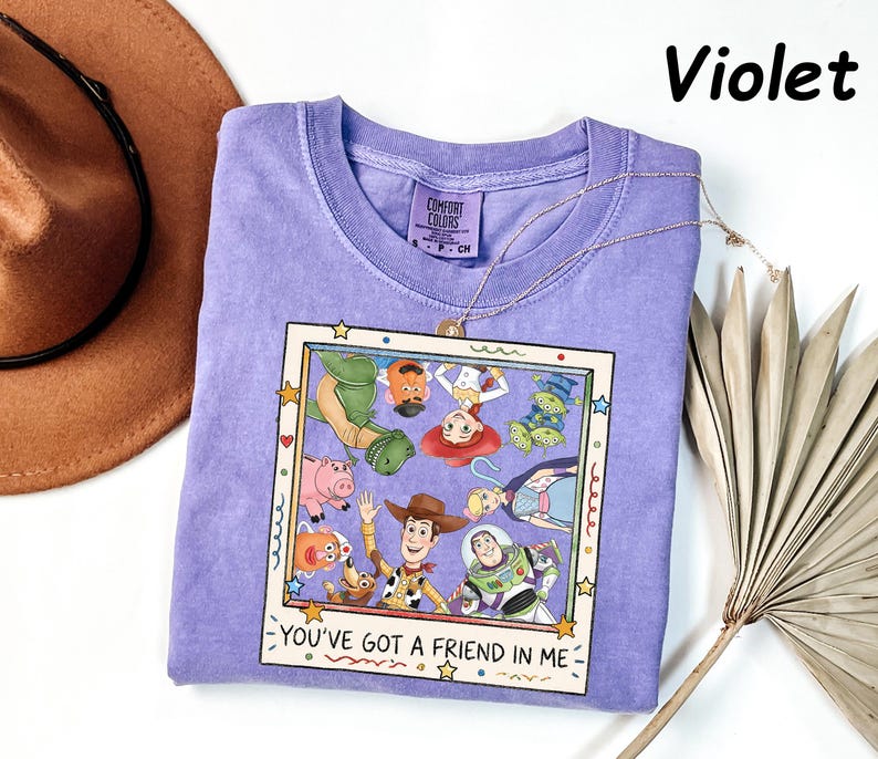 May include: A violet short-sleeved t-shirt featuring a graphic print of Toy Story characters inside a square frame. The text "YOU'VE GOT A FRIEND IN ME" is printed below the frame. The shirt has a Comfort Colors brand label at the neck.