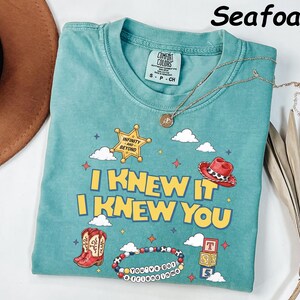 May include: A seafoam green short-sleeve t-shirt featuring a graphic print with the text I KNEW IT I KNEW YOU, a sheriff star with INFINITY AND BEYOND, a red cowboy hat, cowboy boots, a beaded bracelet with YOU'VE GOT A FRIEND IN ME, and blocks.