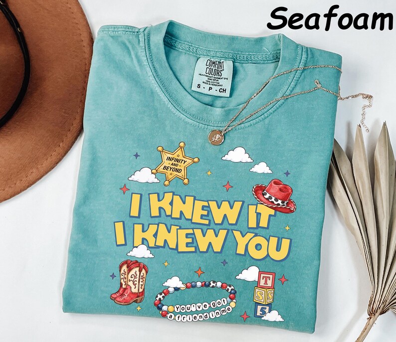 May include: A seafoam green short-sleeve t-shirt featuring a graphic print with the text I KNEW IT I KNEW YOU, a sheriff star with INFINITY AND BEYOND, a red cowboy hat, cowboy boots, a beaded bracelet with YOU'VE GOT A FRIEND IN ME, and blocks.