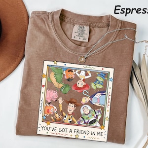 May include: An espresso-colored short-sleeve t-shirt featuring a graphic print of various animated characters inside a square frame. The text "YOU'VE GOT A FRIEND IN ME" is printed below the characters. The shirt has a crew neck and a brand label.