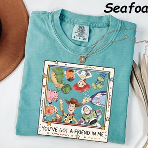 May include: A seafoam green t-shirt featuring a graphic print of various animated characters inside a square frame. The text "YOU'VE GOT A FRIEND IN ME" is printed below the frame. The shirt has a crew neckline and a Comfort Colors brand label.