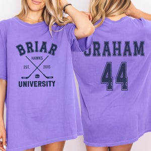 May include: A lilac-colored t-shirt with the text "BRIAR HAWKS UNIVERSITY" and crossed hockey sticks. The back of the shirt displays the name "GRAHAM" and the number "44" in navy blue.