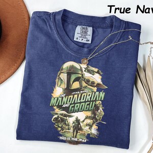 Peut inclure: Un t-shirt bleu marine avec un graphisme de Star Wars repr&eacute;sentant Le Mandalorien et Grogu. Le design comprend le texte "THE STAR WARS MANDALORIAN AND GROGU" et "THIS IS THE WAY". Un collier dor&eacute; est drap&eacute; sur le t-shirt.