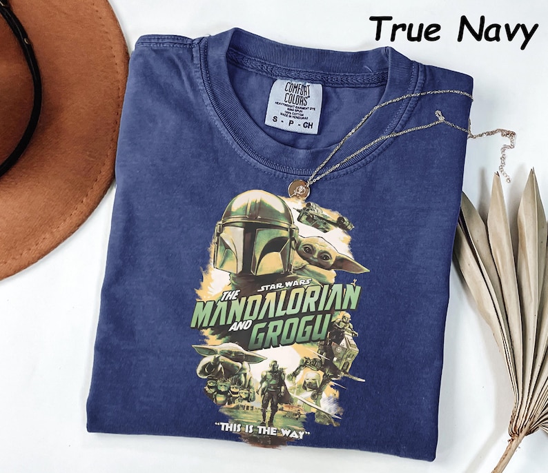 Peut inclure: Un t-shirt bleu marine avec un graphisme de Star Wars repr&eacute;sentant Le Mandalorien et Grogu. Le design comprend le texte "THE STAR WARS MANDALORIAN AND GROGU" et "THIS IS THE WAY". Un collier dor&eacute; est drap&eacute; sur le t-shirt.