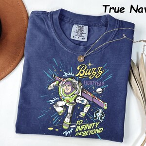 May include: True navy blue t-shirt featuring a graphic of Buzz Lightyear with the text "Buzz Lightyear" and "To Infinity and Beyond." The shirt has a crew neck and short sleeves. A gold necklace with a pendant is draped across the shirt.