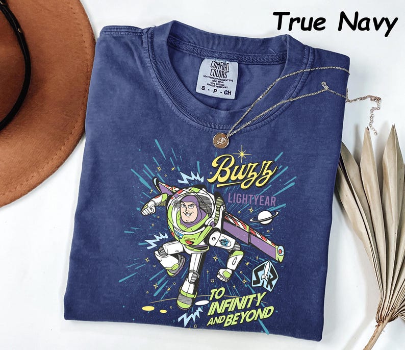 May include: True navy blue t-shirt featuring a graphic of Buzz Lightyear with the text "Buzz Lightyear" and "To Infinity and Beyond." The shirt has a crew neck and short sleeves. A gold necklace with a pendant is draped across the shirt.