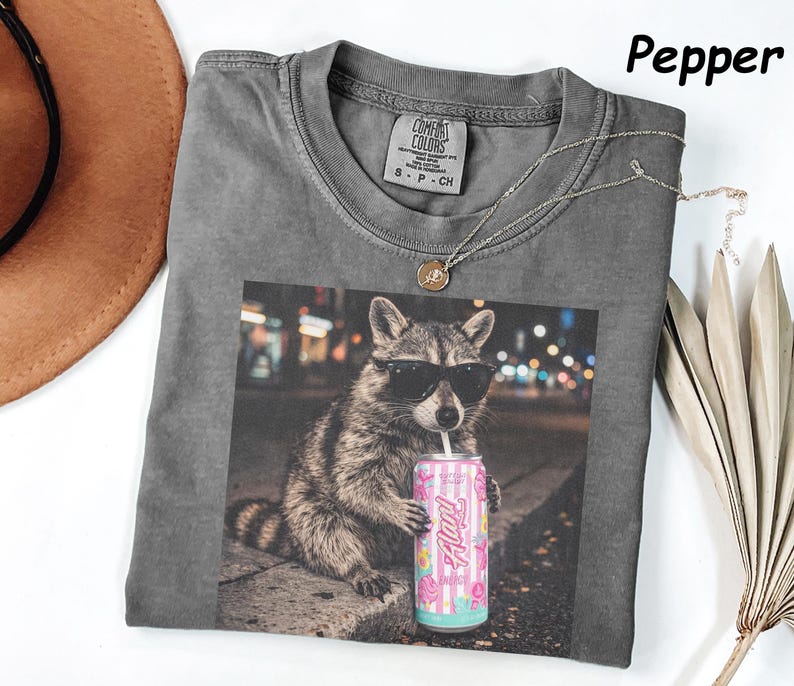 May include: A gray Comfort Colors t-shirt featuring a graphic of a raccoon wearing sunglasses and holding a pink can of "Alani" drink. The shirt has a crew neck and short sleeves. A silver necklace is draped across the shirt.