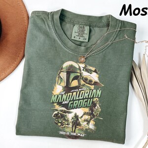 Peut inclure: Un t-shirt vert mousse avec un graphisme de "The Mandalorian" avec le texte "The Mandalorian and Grogu" et "This is the Way". Un collier dor&eacute; est drap&eacute; sur le t-shirt. Le t-shirt est pli&eacute; sur une surface blanche.