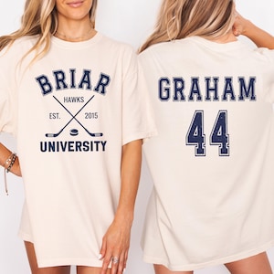 May include: Cream-colored t-shirt featuring the text "BRIAR HAWKS UNIVERSITY" with crossed hockey sticks and the year 2015. The back of the shirt displays "GRAHAM 44" in navy blue lettering.
