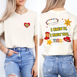 May include: A cream-colored t-shirt featuring a small red heart with yellow TS letters on the front. The back displays a colorful graphic with a beaded bracelet, a sheriff star, red cowboy boots, a red hat, and the text I KNEW IT, I KNEW YOU.