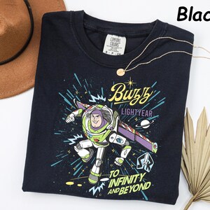 May include: Black crew neck t-shirt featuring a Buzz Lightyear graphic. The design includes the text "Buzz Lightyear" and "To Infinity and Beyond". A gold necklace and a brown hat are also visible.