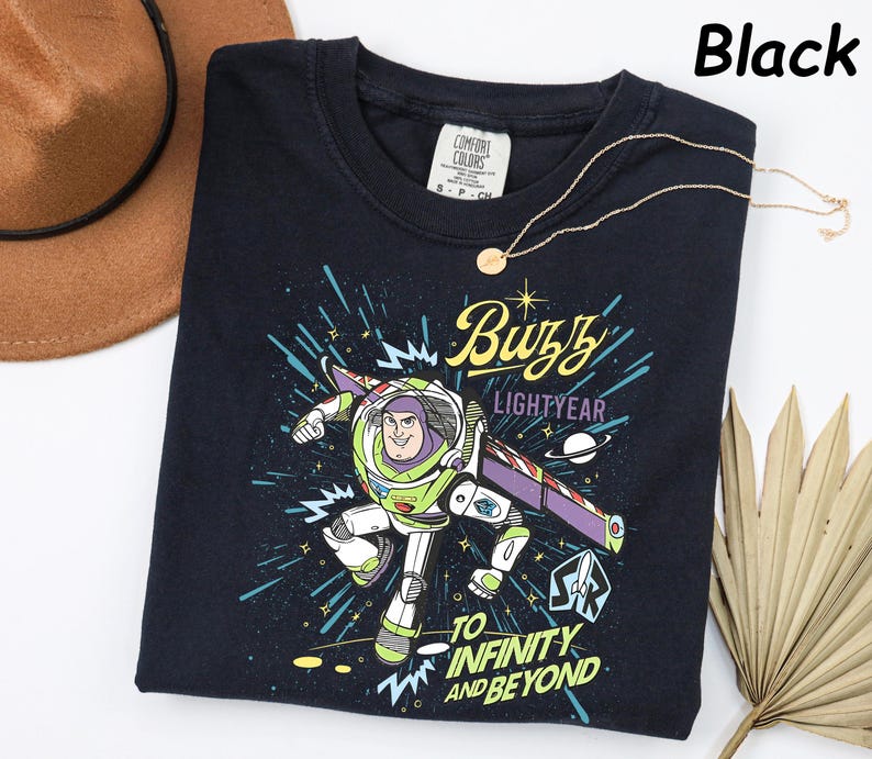 May include: Black crew neck t-shirt featuring a Buzz Lightyear graphic. The design includes the text "Buzz Lightyear" and "To Infinity and Beyond". A gold necklace and a brown hat are also visible.