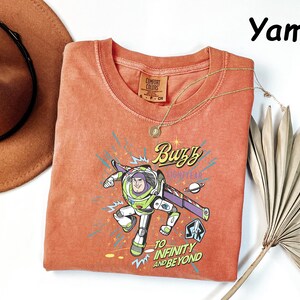 May include: Orange-red Buzz Lightyear t-shirt with a graphic of the character and the phrase "To Infinity and Beyond." The shirt has a crew neck and short sleeves. A gold necklace and a brown hat are also in the image.