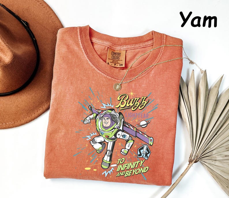 May include: Orange-red Buzz Lightyear t-shirt with a graphic of the character and the phrase "To Infinity and Beyond." The shirt has a crew neck and short sleeves. A gold necklace and a brown hat are also in the image.