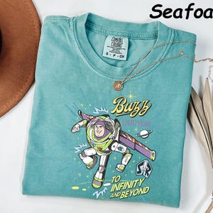 May include: Seafoam green t-shirt featuring a graphic of Buzz Lightyear with the text "Buzz Lightyear" and "To Infinity and Beyond." The shirt has a crew neck and short sleeves. A gold necklace with a pendant is draped across the shirt.