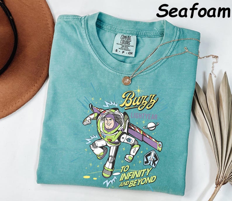 May include: Seafoam green t-shirt featuring a graphic of Buzz Lightyear with the text "Buzz Lightyear" and "To Infinity and Beyond." The shirt has a crew neck and short sleeves. A gold necklace with a pendant is draped across the shirt.
