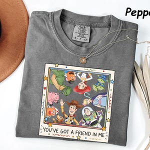 May include: A pepper-colored short-sleeve t-shirt featuring a graphic print of Toy Story characters inside a square frame. The design includes Woody, Buzz Lightyear, Jessie, and others with the text "YOU'VE GOT A FRIEND IN ME" at the bottom.