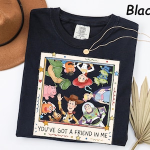 May include: A black t-shirt featuring a square graphic print of various animated characters from Toy Story arranged in a frame. The text "YOU'VE GOT A FRIEND IN ME" is printed at the bottom of the frame. The shirt has a white neck label.