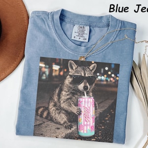 May include: Blue jean colored t-shirt with a graphic of a raccoon wearing sunglasses and holding a can of soda. The can has a floral design and the word "Flirty". The shirt has a crew neck and a small tag.