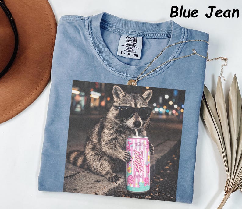 May include: Blue jean colored t-shirt with a graphic of a raccoon wearing sunglasses and holding a can of soda. The can has a floral design and the word "Flirty". The shirt has a crew neck and a small tag.