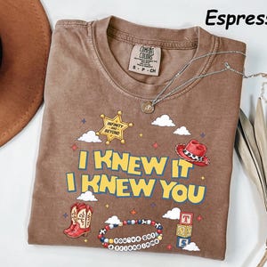 May include: An espresso-colored t-shirt featuring a graphic print with the text I KNEW IT I KNEW YOU. The design includes a yellow sheriff star with INFINITY AND BEYOND, a red cowboy hat, red boots, a beaded bracelet with YOU'VE GOT A FRIEND IN ME.