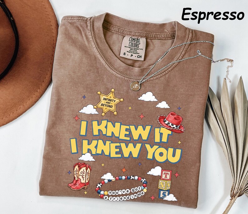 May include: An espresso-colored t-shirt featuring a graphic print with the text I KNEW IT I KNEW YOU. The design includes a yellow sheriff star with INFINITY AND BEYOND, a red cowboy hat, red boots, a beaded bracelet with YOU'VE GOT A FRIEND IN ME.