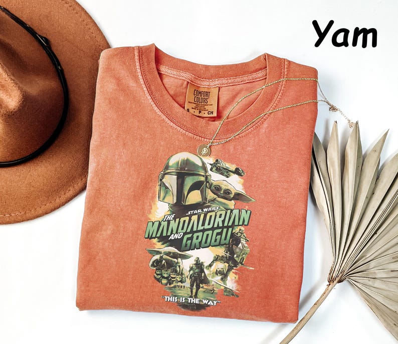 Peut inclure: Un t-shirt col rond de couleur corail avec un graphisme de "The Mandalorian and Grogu". Le motif comprend le casque du Mandalorien, Grogu, et le texte "The Mandalorian and Grogu" et "This is the Way."