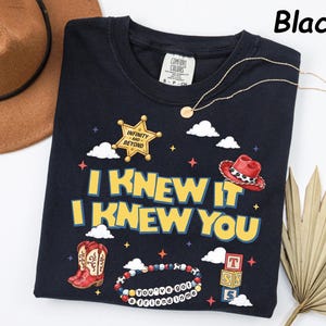 May include: A black t-shirt featuring a graphic print with the text "I KNEW IT I KNEW YOU" in yellow letters. The design includes a gold sheriff badge with "INFINITY AND BEYOND", a red cowboy hat, red cowboy boots, a beaded bracelet, and blocks.