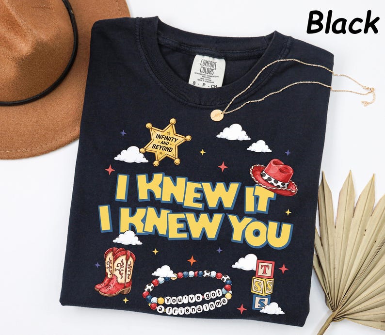 May include: A black t-shirt featuring a graphic print with the text "I KNEW IT I KNEW YOU" in yellow letters. The design includes a gold sheriff badge with "INFINITY AND BEYOND", a red cowboy hat, red cowboy boots, a beaded bracelet, and blocks.