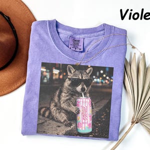 May include: Violet-colored t-shirt featuring a graphic of a raccoon wearing sunglasses and drinking from a can. The can has a pink and white striped design with the word "Alani" on it. The shirt has a crew neck and short sleeves.
