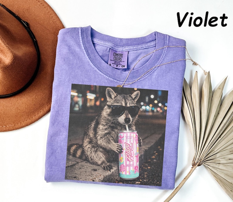 May include: Violet-colored t-shirt featuring a graphic of a raccoon wearing sunglasses and drinking from a can. The can has a pink and white striped design with the word "Alani" on it. The shirt has a crew neck and short sleeves.
