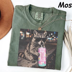 May include: Moss green t-shirt with a graphic of a raccoon wearing sunglasses and holding a pink can. The shirt has a crew neck and short sleeves. A gold necklace is draped across the shirt.