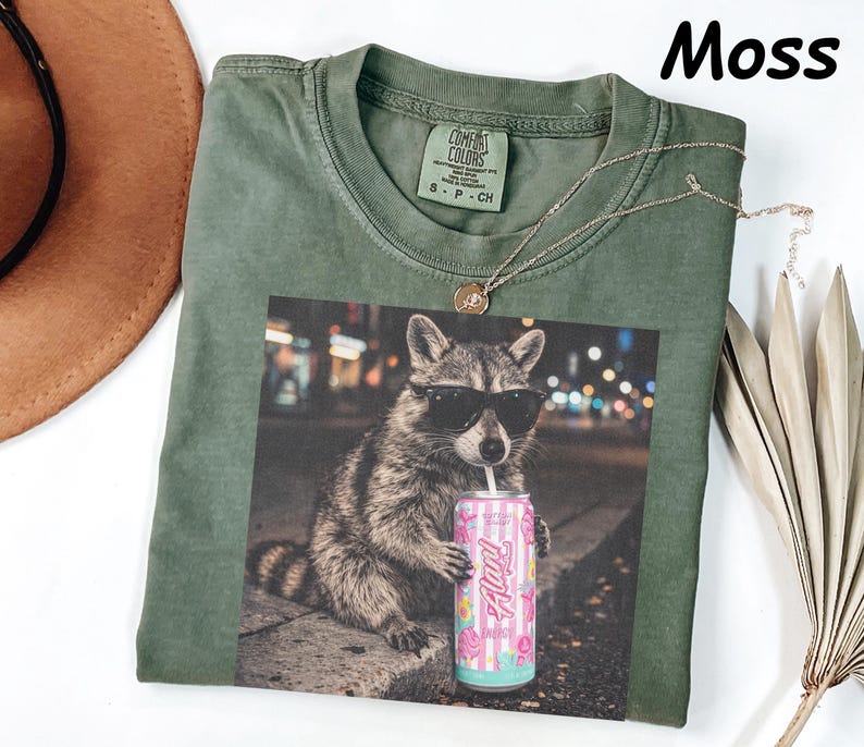 May include: Moss green t-shirt with a graphic of a raccoon wearing sunglasses and holding a pink can. The shirt has a crew neck and short sleeves. A gold necklace is draped across the shirt.