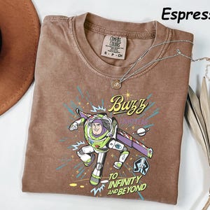 May include: Espresso-colored t-shirt featuring a graphic of Buzz Lightyear with the text "Buzz Lightyear" and "To Infinity and Beyond." The shirt has a crew neck and a small tag on the upper left. A silver necklace with a pendant is draped across the shirt.