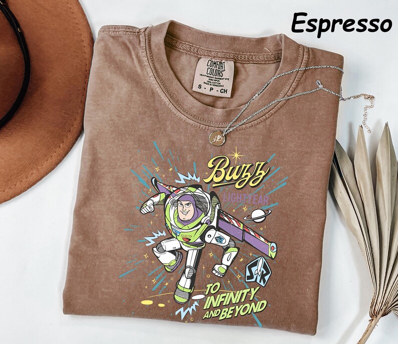 May include: Espresso-colored t-shirt featuring a graphic of Buzz Lightyear with the text "Buzz Lightyear" and "To Infinity and Beyond." The shirt has a crew neck and a small tag on the upper left. A silver necklace with a pendant is draped across the shirt.