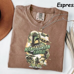 Peut inclure: T-shirt couleur expresso avec un graphisme de The Mandalorian et Grogu de Star Wars. Le t-shirt a un col rond et des manches courtes. Un collier argent&eacute; avec un pendentif repose sur le t-shirt. Le texte "THIS IS THE WAY" est imprim&eacute;.