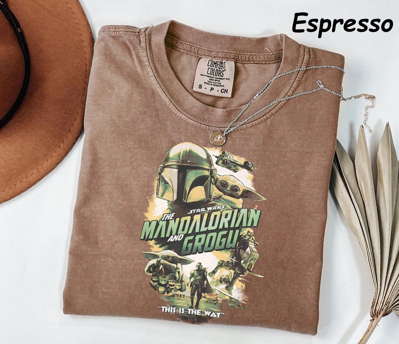 Peut inclure: T-shirt couleur expresso avec un graphisme de The Mandalorian et Grogu de Star Wars. Le t-shirt a un col rond et des manches courtes. Un collier argent&eacute; avec un pendentif repose sur le t-shirt. Le texte "THIS IS THE WAY" est imprim&eacute;.