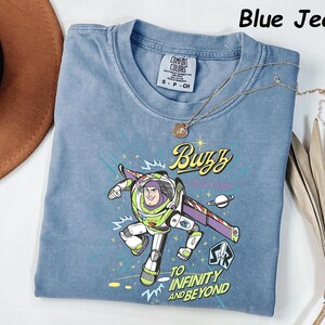 May include: Blue jean colored t-shirt featuring a graphic of Buzz Lightyear with the text "Buzz" and "To Infinity and Beyond". The shirt has a crew neck and short sleeves. A gold necklace with a pendant is draped across the shirt.