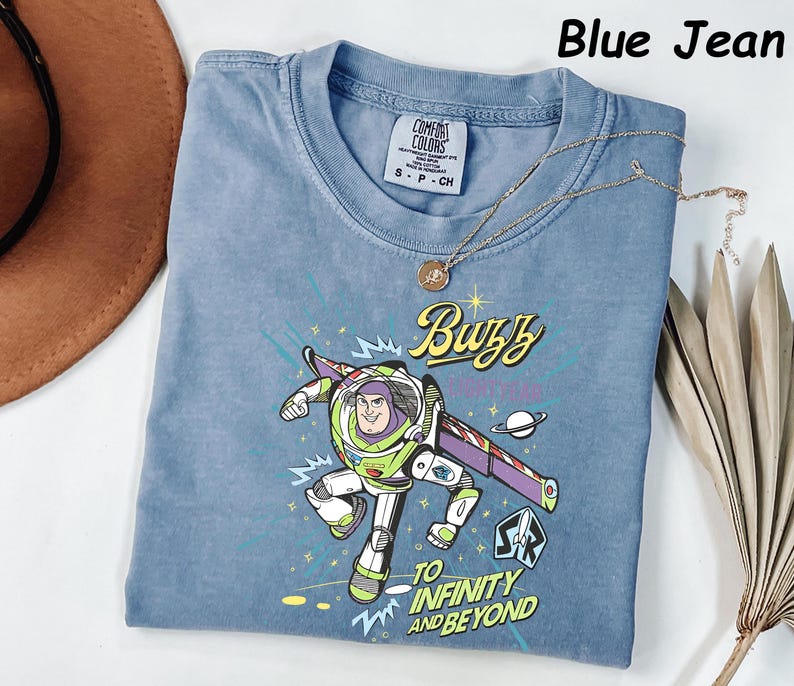 May include: Blue jean colored t-shirt featuring a graphic of Buzz Lightyear with the text "Buzz" and "To Infinity and Beyond". The shirt has a crew neck and short sleeves. A gold necklace with a pendant is draped across the shirt.