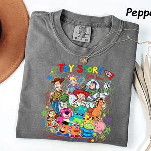 May include: A gray t-shirt featuring a colorful graphic of characters from the animated film "Toy Story." The design includes Woody, Buzz Lightyear, and other characters, with the words "Toy Story" and "yee haw" in a playful font. The shirt is a Comfort Colors brand.