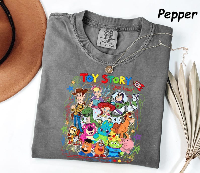 May include: A gray t-shirt featuring a colorful graphic of characters from the animated film "Toy Story." The design includes Woody, Buzz Lightyear, and other characters, with the words "Toy Story" and "yee haw" in a playful font. The shirt is a Comfort Colors brand.