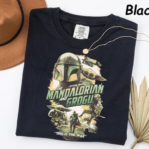 Peut inclure: T-shirt noir avec un graphisme de "The Mandalorian" avec le texte "The Mandalorian and Grogu" et "This is the Way". Le design comprend des images du Mandalorien et de Grogu. Un collier en or est drap&eacute; sur le t-shirt.