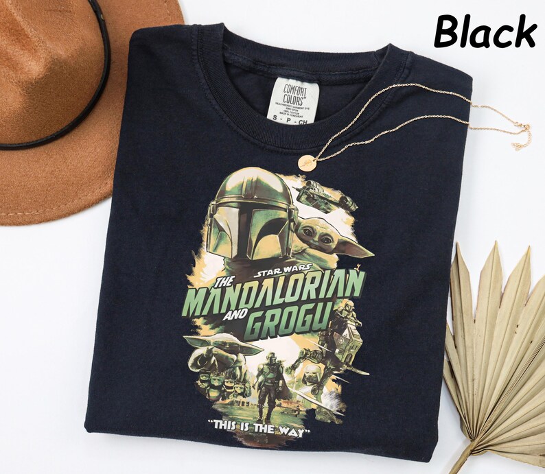 Peut inclure: T-shirt noir avec un graphisme de "The Mandalorian" avec le texte "The Mandalorian and Grogu" et "This is the Way". Le design comprend des images du Mandalorien et de Grogu. Un collier en or est drap&eacute; sur le t-shirt.