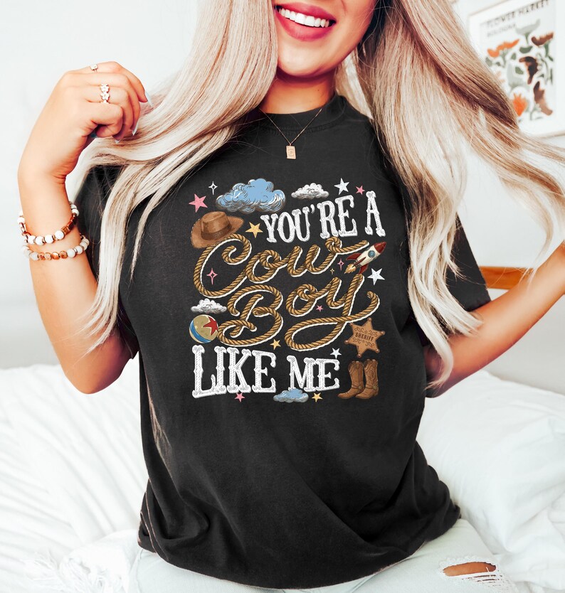 K&ouml;nnte beinhalten: Ein schwarzes T-Shirt mit einem Grafikdruck und dem Text "YOU'RE A COW BOY LIKE ME" in einer Schriftart, die wie ein Seil aussieht. Das Design enth&auml;lt Illustrationen eines braunen Cowboyhuts, einer Rakete, eines Balls, eines Sheriffsterns und brauner Stiefel.