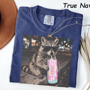 May include: Navy blue t-shirt with a graphic of a raccoon wearing sunglasses and holding a pink and blue can of "Alani Nu" drink. The shirt has a Comfort Colors tag and the words "True Navy" are printed at the top.
