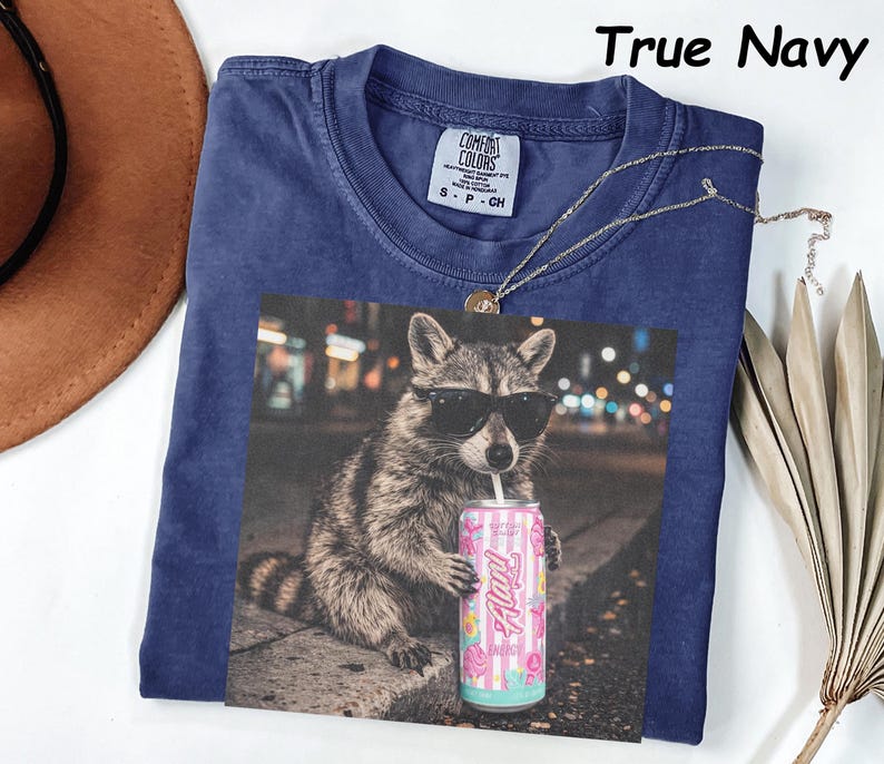 May include: Navy blue t-shirt with a graphic of a raccoon wearing sunglasses and holding a pink and blue can of "Alani Nu" drink. The shirt has a Comfort Colors tag and the words "True Navy" are printed at the top.