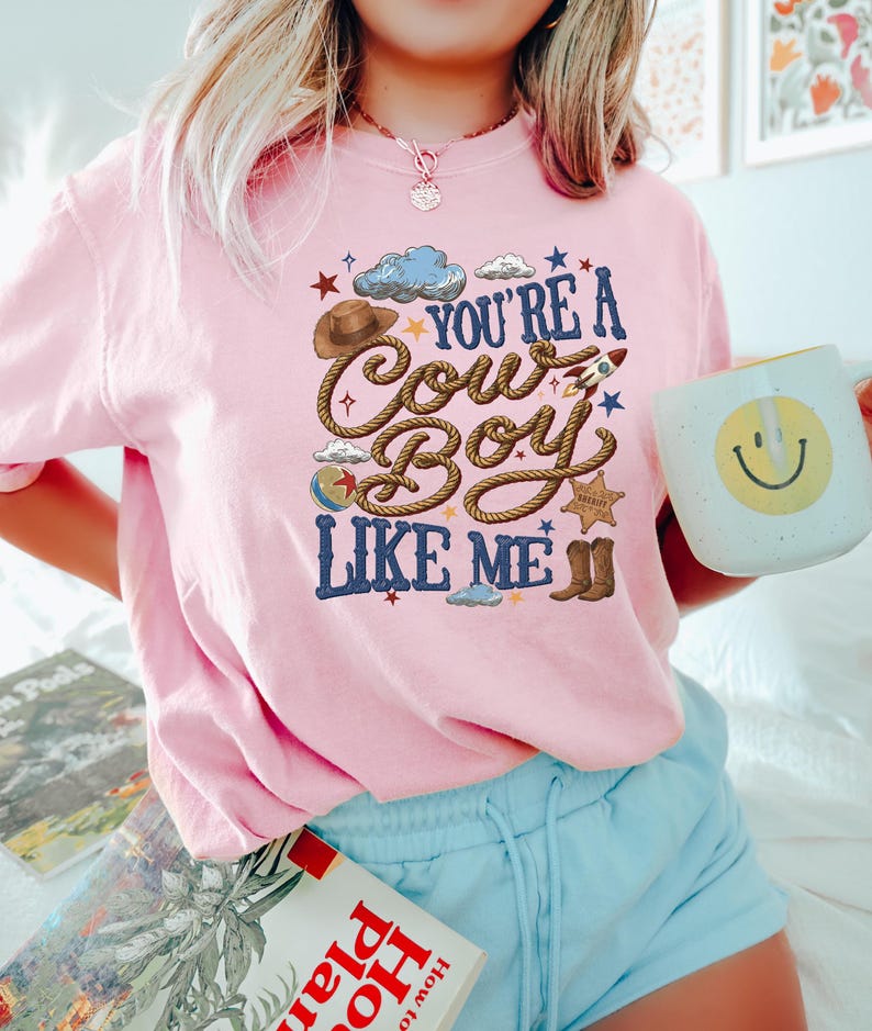 K&ouml;nnte beinhalten: Ein hellrosa T-Shirt mit einem Grafikdruck und dem Text "YOU'RE A COWBOY LIKE ME" in blauen Buchstaben. Das Design zeigt einen braunen Cowboyhut, ein Seil, eine kleine Rakete, einen Sheriffstern, braune Stiefel, einen Ball und blaue Wolken.