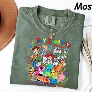 May include: Moss green t-shirt featuring a colorful graphic of Toy Story characters. The design includes Woody, Buzz Lightyear, and other beloved characters from the animated film. The words "Toy Story" and "yee haw" are also visible.